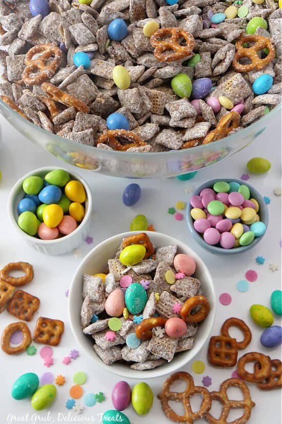 A white surface with small bowls filled with pastel candies and one bowl filled with a muddy buddy mix with an Easter theme.