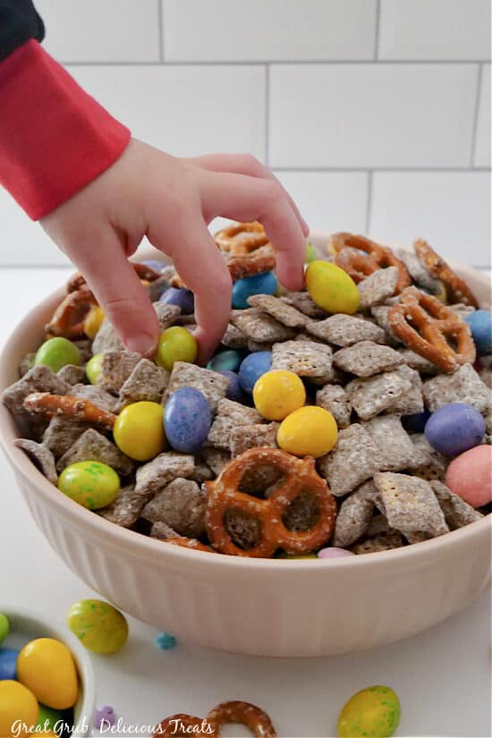 A light pink bowl filled with muddy buddies and a toddler's fingers picking out a yellow chocolate candy.