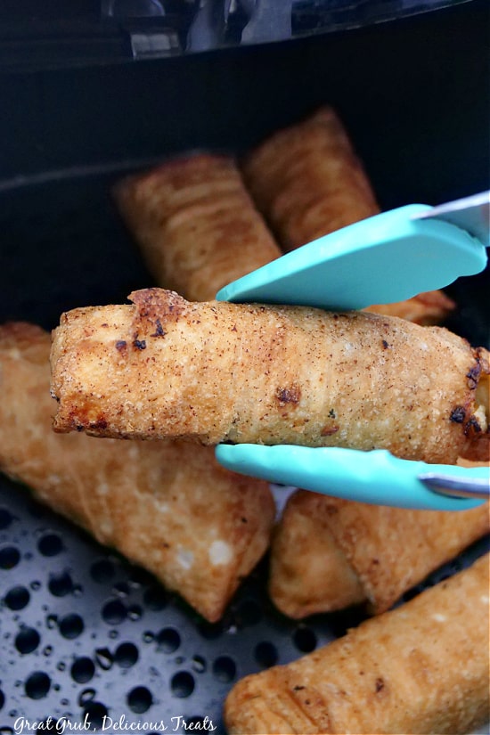 A pair of tongs lifting an egg roll out of the air fryer basket.