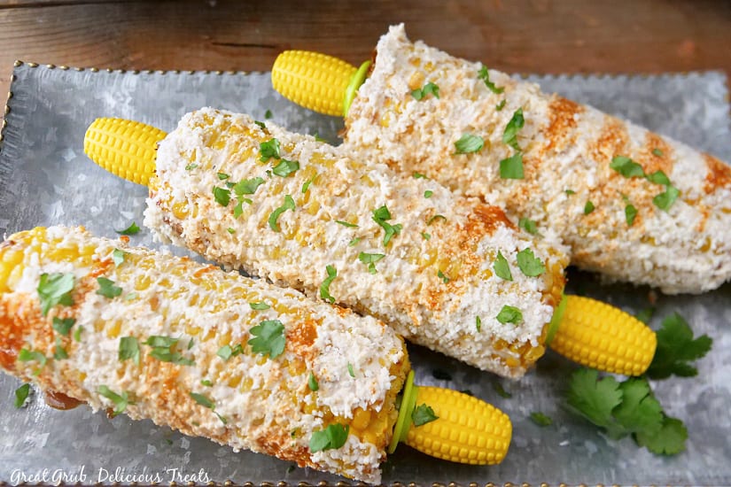 A silver tray with three corn on the cobs covered in a delicious mix of elote toppings.