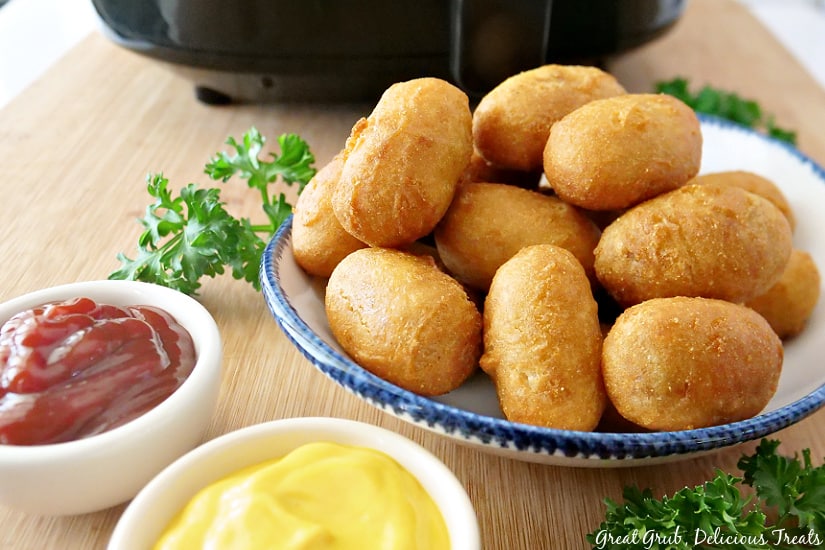 a white plate with blue trim loaded with mini corn dogs, a small bowl full of ketchup and another small bowl of mustard. 