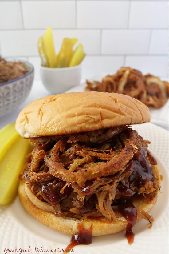 A pulled pork sandwich placed on a white plate with two pickles next to it.