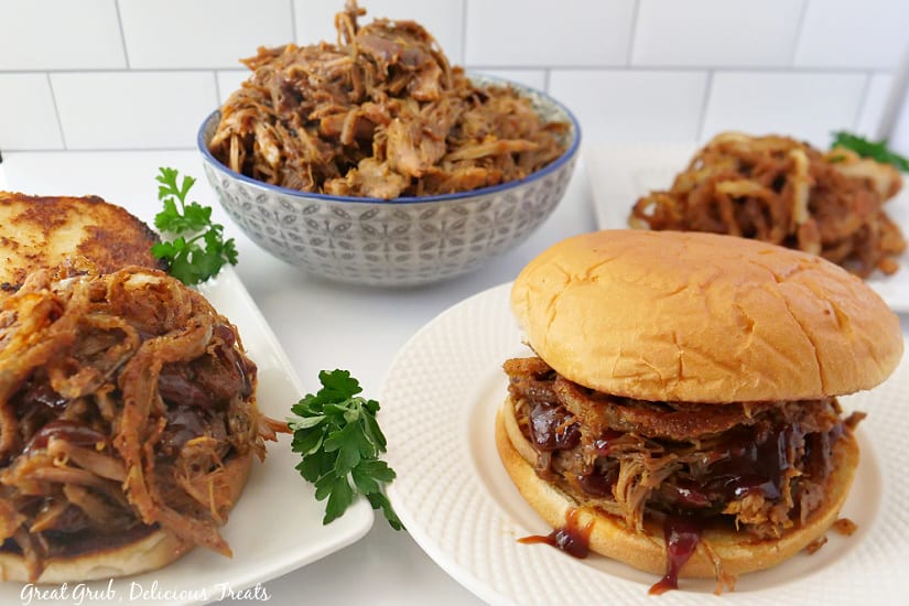 A horizontal picture of BBQ pulled pork sandwiches on white plates with a bowl full of pulled pork in the background.