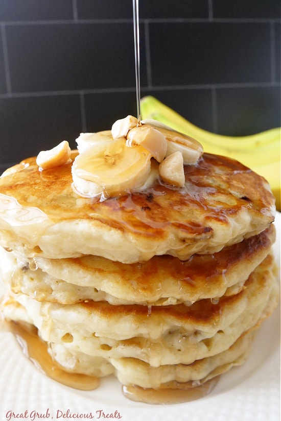 Three pancakes stacked up on a white plate with sliced bananas and macadamia nuts on top with a banana in the background for decoration.