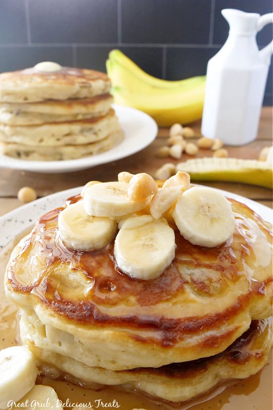 Pancakes stacked on a white plate with maple syrup and sliced bananas and macadamia nuts.