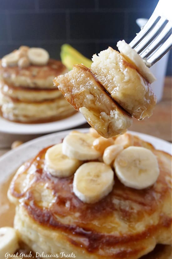 A bite of pancakes on a fork, with two white plates with pancakes stacked on them in the background.