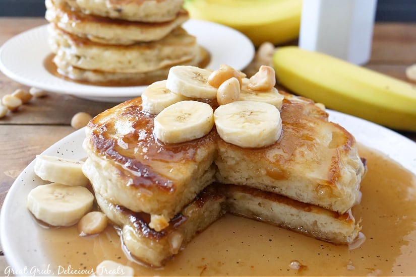 A large white plate with two banana pancakes with maple syrup, topped with sliced bananas and macadamia nuts with two bananas in the background.