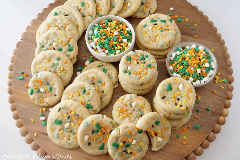 A round wooden surface with two dozen cookies on it with two small white bowls filled with a baking chips, mini marshmallows and sprinkles.