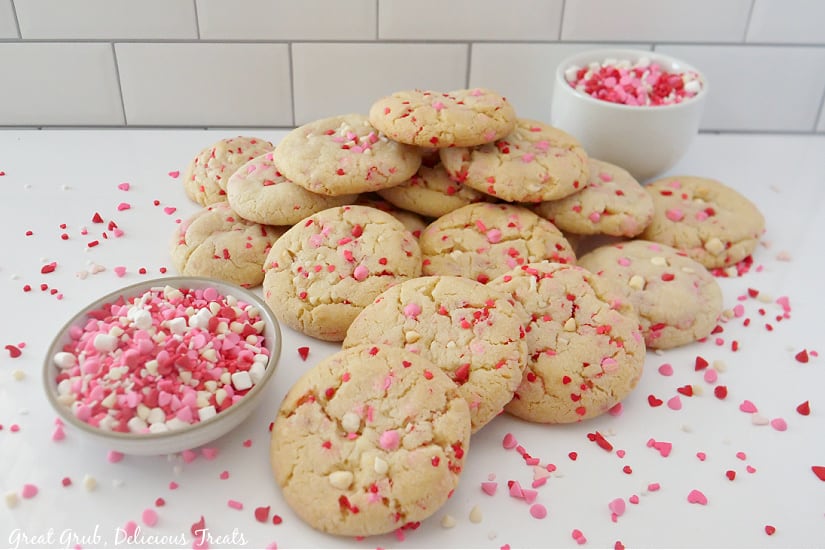 A horizontal picture of Valentine's Day sugar cookies stacked in a pile with sprinkles spread around the cookies.