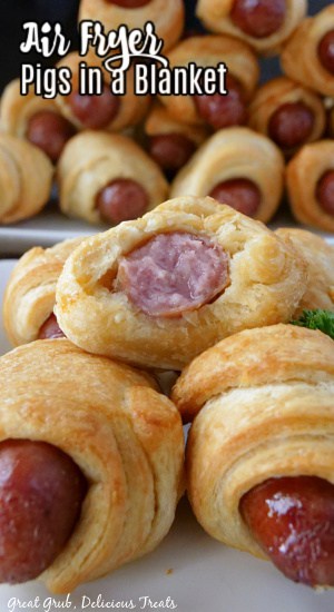 Pigs in a blanket stacked up on a plate with a large plate  in the background filled with crescent rolls.