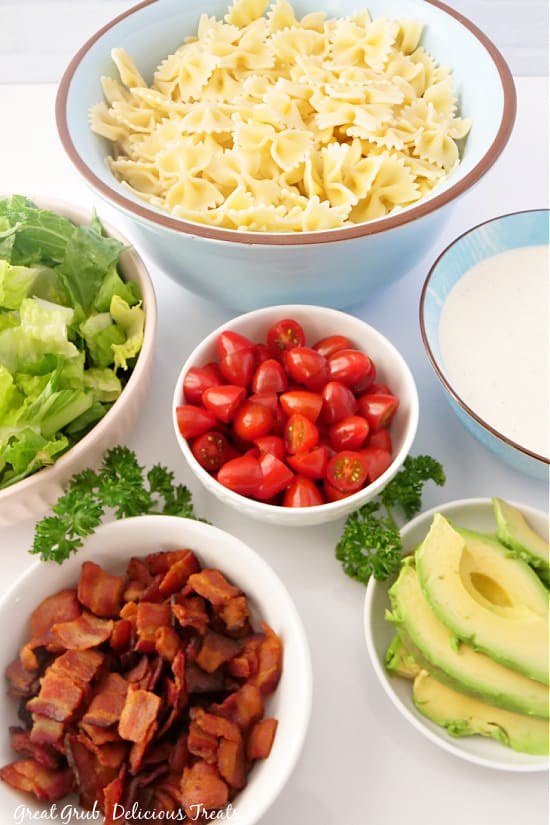 Bowls filled with each ingredients of this recipe; sliced avocados in a white bowl, diced grape tomatoes in a white bowl, lettuce in a white bowl, chopped crispy bacon in a white bowl, bow tie pasta noodles in a large bowl, and a blue bowl full of ranch dressing.