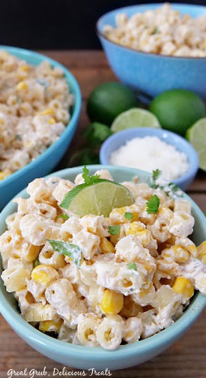 A small bowl filled with Mexican street corn pasta salad, a small bowl of cojita cheese, and limes in the background.