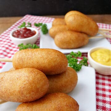 Two white plates with mini corn dogs on them and two small bowls of ketchup and mustard.