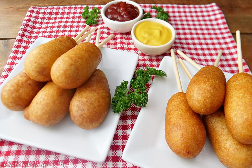 Two white, square plates with corn dogs on them, sitting on a red and white place mat, with dipped sauce in the background.