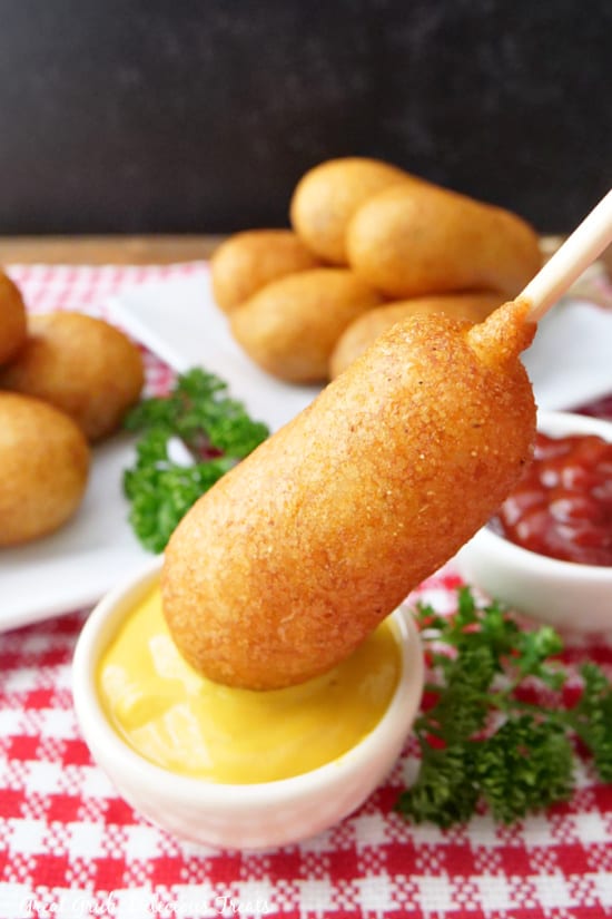 A corn dog on a stick being dipped in mustard.
