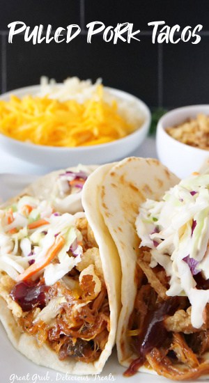 Two tacos on a plate with shredded cabbage and onions strings on top. A small white bowl of shredded cheese in the background.