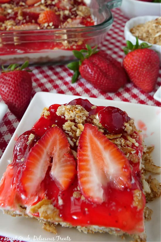 An overhead shot of a piece of strawberry dessert on a white plate with a red and white checkered placemat underneath and a baking dish with dessert in the background.