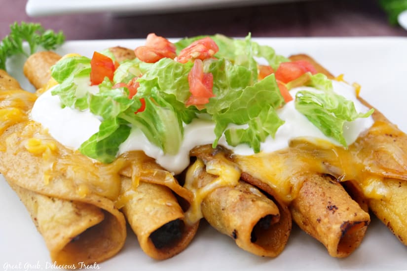 Five taquitos on a white plate, topped with melted cheese, sour cream, and lettuce.