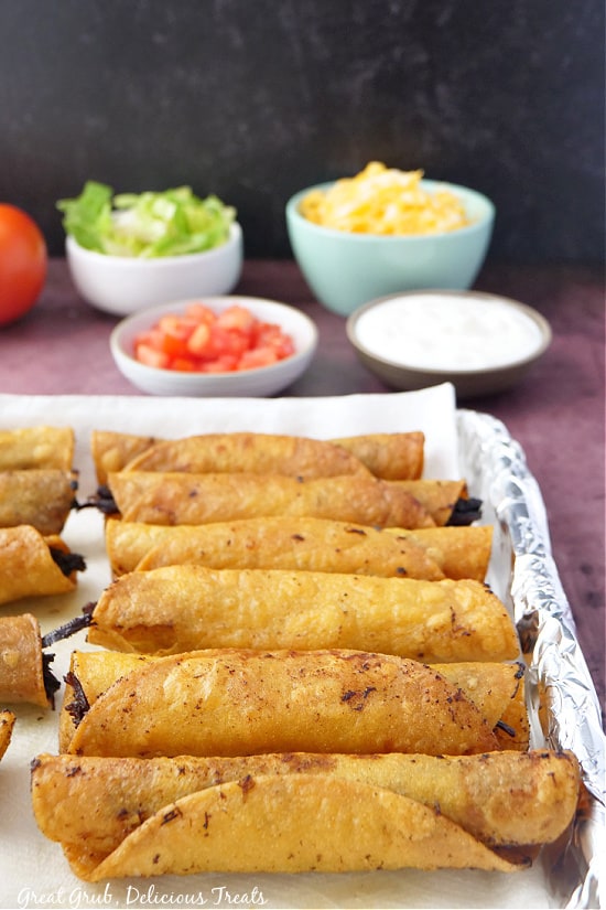 A cookie sheet with foil, with freshly fried taquitos on it, with all the toppings in the background.