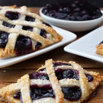 Individual pastries on white square plates with a bowl of blueberry pie filling in the background.