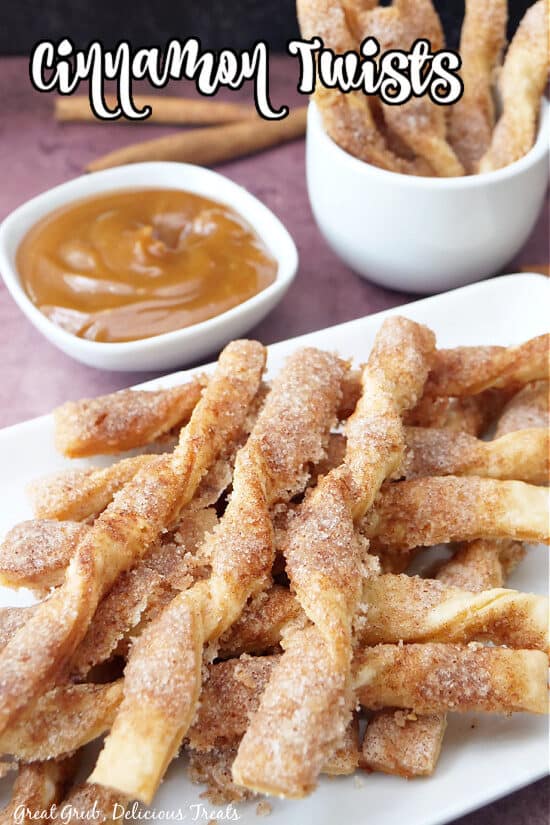 A close-up picture of cinnamon twists stacked on a white plate with a small bowl of caramel dipping sauce and more twists in the background.