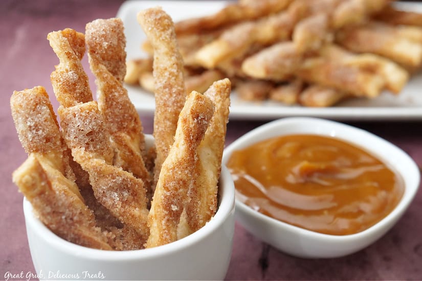 A horizontal picture of Cinnamon twists standing upright in a white container with a small bowl of caramel dipping sauce next to it and more Cinnamon Twists in the background.