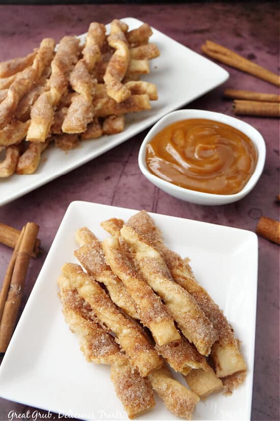 An overhead picture of two plates with Cinnamon Twists stacked on them with a small white bowl filled with caramel sauce, and cinnamon sticks all around.