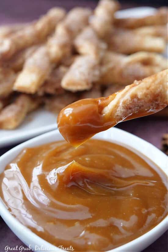 A picture of a white bowl filled with caramel sauce and a cinnamon twist dipped in caramel with a plate stacked with cinnamon twists in the background.