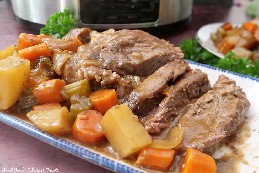 A horizontal photo of a chuck roast and vegetables on a white rectangle plate with blue trim.