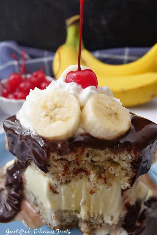 A slice of cake on a plate with a bite taken out of it, topped with hot fudge, sliced bananas, whipped cream, and a cherry.