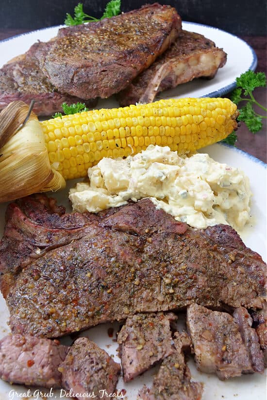 Two plates with steak on them, served with corn on the cob and potato salad.