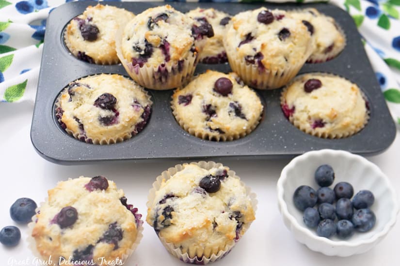 A landscape photo of a muffin tin with freshly baked muffins and a small white bowl with blueberries.