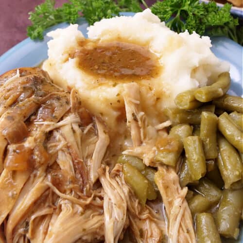 A serving of Mississippi chicken on a blue plate with mashed potatoes, green beans, and gravy.