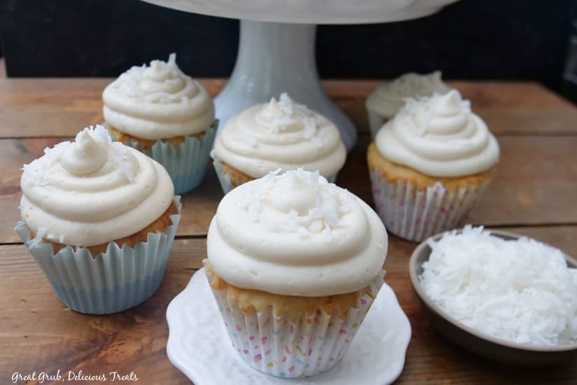 A landscape photo of seven cupcakes sitting on a wooden board, with a small bowl of shredded coconut in the background.