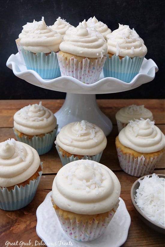 Coconut cupcakes topped with frosting and shredded coconut sitting on cake display plates and on a wooden board.