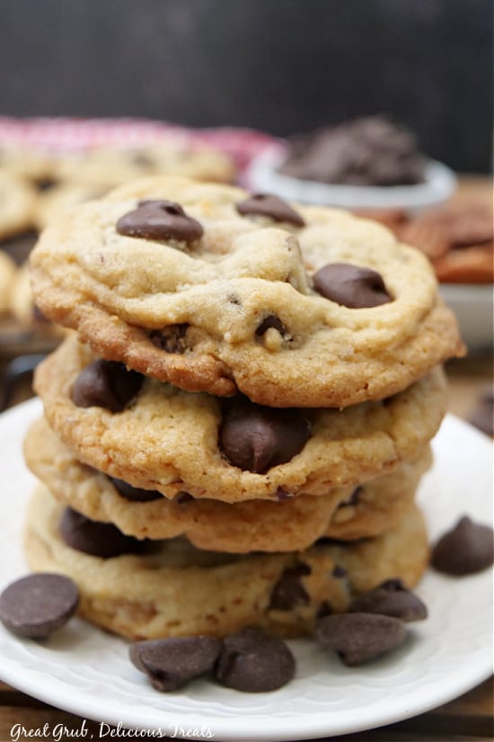 A stack of four chocolate chip cookies on a white plate with chocolate chips sprinkled in the front. 