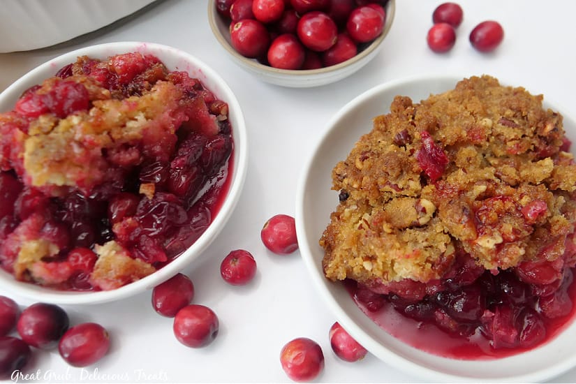 Two white bowl full of cobbler, a bowl of cranberries, and cranberries scattered around the bowls.