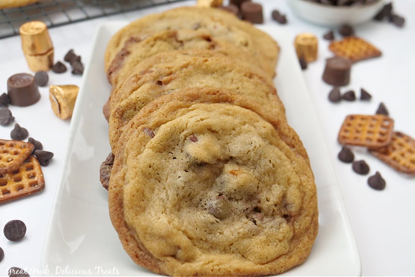 Cookies lined up on a long white plate with pretzels, Rolos, and chocolate chips laying around the background.