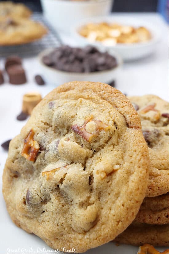 A stack of cookies on a white plate with two small bowls in the background filled with chocolate chips and Rolos. 