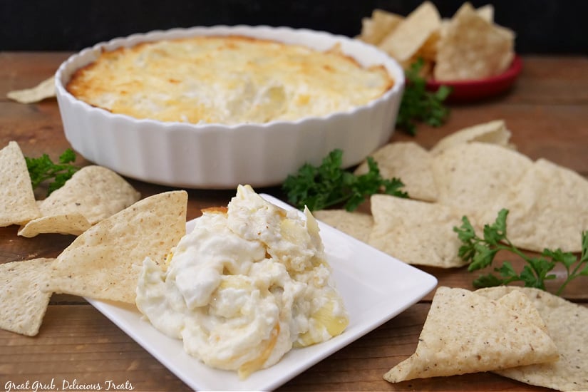 A horizontal photo of a white plate with artichoke dip on it with a round white bowl in the background and tortilla chips spread around.
