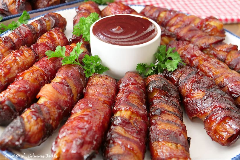 A horizontal photo of a white plate with stuffed manicotti shells wrapped in bacon placed around a small bowl of barbecue sauce.