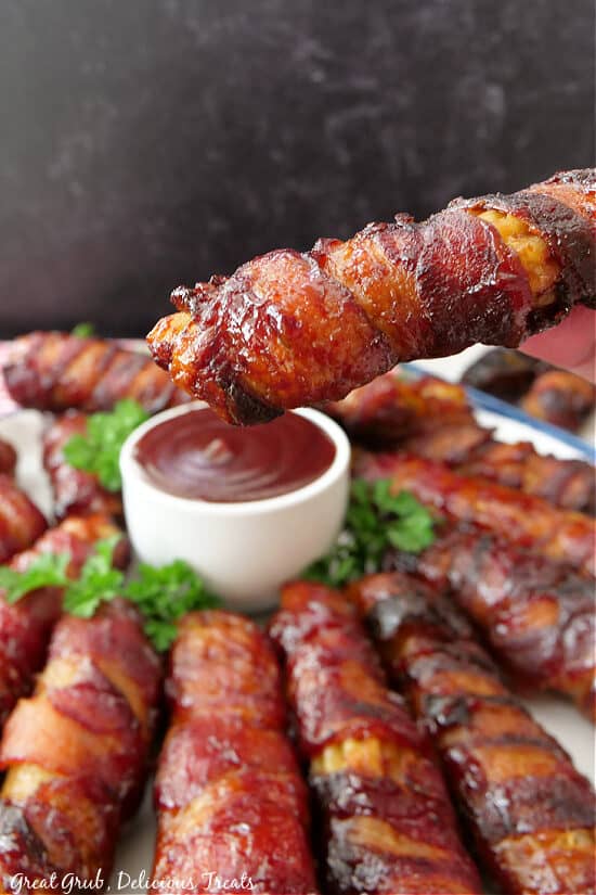 A bacon wrapped stuffed manicotti shell after being smoked held above a small bowl of barbecue sauce.