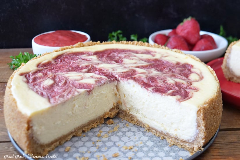 A horizontal photo of a cheesecake with a few slices missing with strawberries and strawberry sauce in the background.