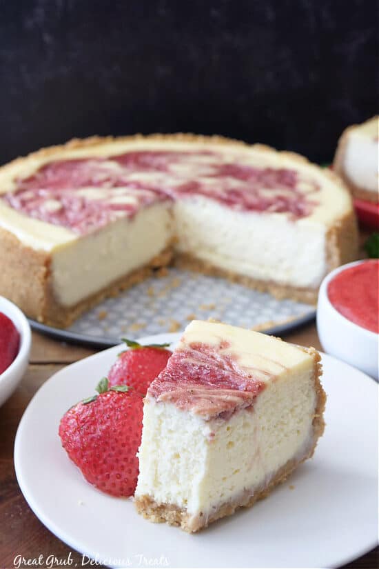 A slice of cheesecake on a white plate with the whole cheesecake in the background.