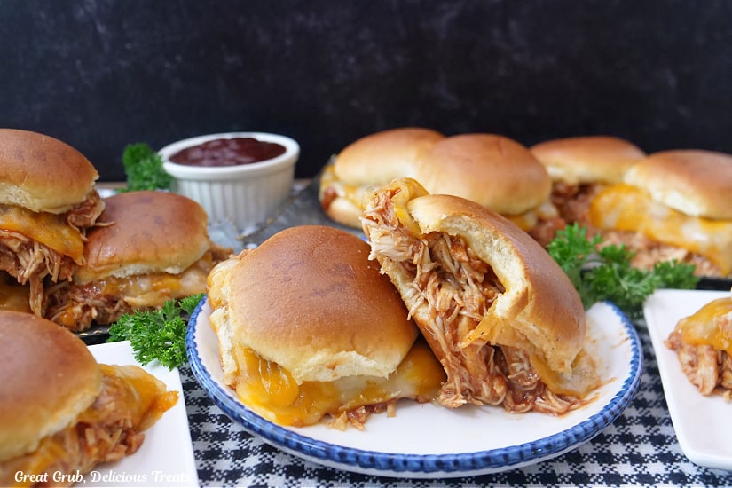 A horizontal photo of barbecue chicken sliders on white plates with more in the background all on a black and white checkered surface.