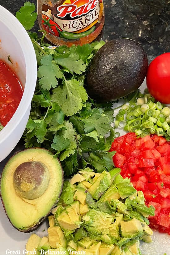 Diced avocado, diced tomatoes, sliced green onions and cilantro on a white surface before being place in a white bowl with picante sauce in it.