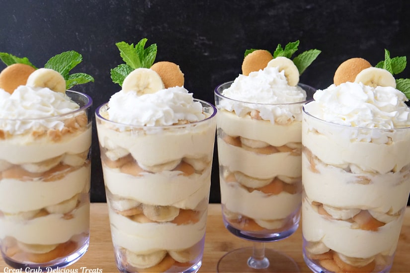 A horizontal photo of four tall parfait glasses filled with banana pudding, banana slices and vanilla wafers, topped with whipped cream.