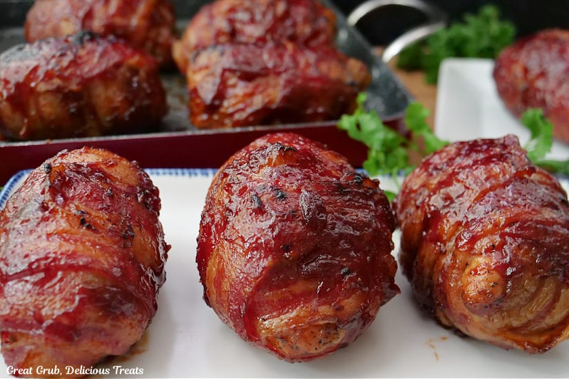 A white plate with blue trim with three bacon wrapped chicken bombs on it with more in the background.