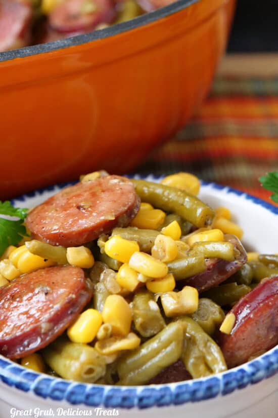 A white bowl with blue trim with a serving of corn, green beans and sliced sausage in it with an orange cast iron pan in the background.