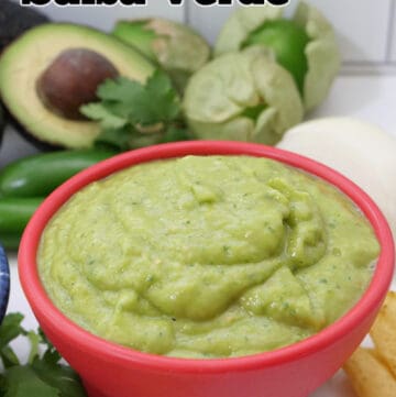 A red bowl filled with creamy avocado salsa verde with the ingredients used placed in the background.